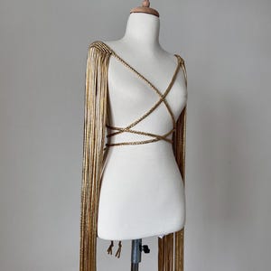 Puede incluir: Joyería corporal dorada con mangas largas con flecos y un diseño entrecruzado. La prenda se exhibe en un maniquí blanco sobre un fondo neutro. La joyería está hecha de material trenzado.
