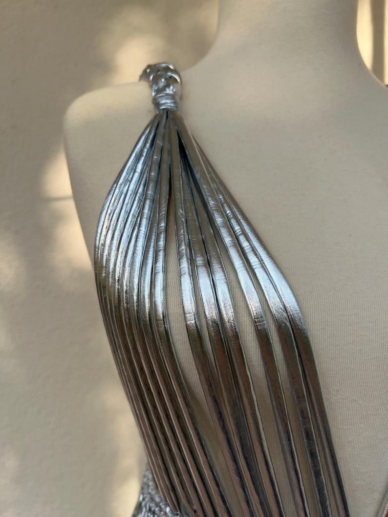 May include: A silver dress with a unique strap design. The strap is made of multiple thin, metallic strips that cascade down the shoulder. The dress is displayed on a mannequin, showcasing the intricate details and texture of the fabric.