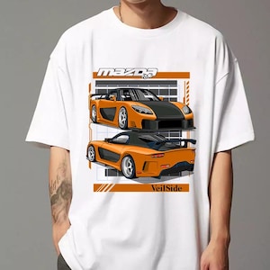 Pu&ograve; includere: Maglietta bianca con una stampa grafica di due auto sportive Mazda RX-7 arancioni. Il testo "mazda RX-7" e "VeilSide" &egrave; stampato sulla maglietta.