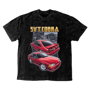 May include: Black t-shirt featuring a graphic of two red SVT Cobra cars. The cars are depicted in a stylized illustration with a gold "SVT COBRA" text at the top. The t-shirt has a vintage, acid wash appearance.