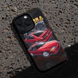 May include: Black phone case featuring a red sports car graphic with the word "COBRA" in gold. The design includes a detailed illustration of the car's exterior and interior, set against a dark background.