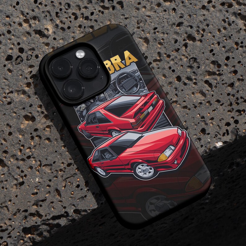 May include: Black phone case featuring a red sports car graphic with the word "COBRA" in gold. The design includes a detailed illustration of the car's exterior and interior, set against a dark background.