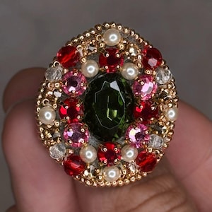 May include: An oval-shaped brooch with a gold-colored beaded border. The center features a large, dark green gemstone, surrounded by red, pink, and clear gemstones, along with small white pearls. The brooch is a vibrant and ornate piece of jewelry.