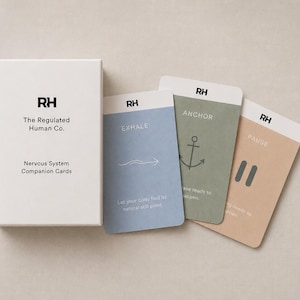 Op de afbeelding: Een witte rechthoekige doos met het label The Regulated Human Co. Nervous System Companion Cards naast drie kaarten. De kaarten zijn lichtblauw met het woord EXHALE, saliegroen met het woord ANCHOR en een ankerpictogram, en beige met het woord PAUSE.