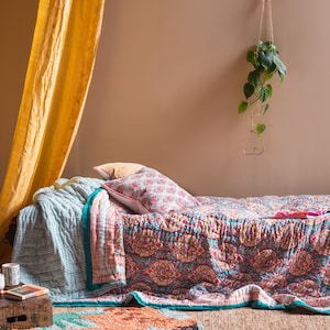 Könnte beinhalten: Ein Bett mit einer türkisfarbenen und rosafarbenen gemusterten Bettdecke und einem weißen und rosafarbenen gemusterten Kissen. Das Bett steht in einem Raum mit einem gelben Vorhang und einer hängenden Pflanze.