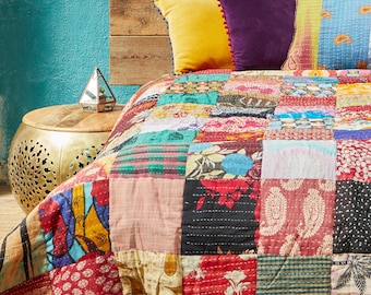 Puede incluir: Una cama con un edredón de retazos en varios colores y patrones. La cama tiene un cabecero de madera y está adornada con almohadas amarillas y moradas. Una mesita auxiliar dorada y un puf estampado están en primer plano.