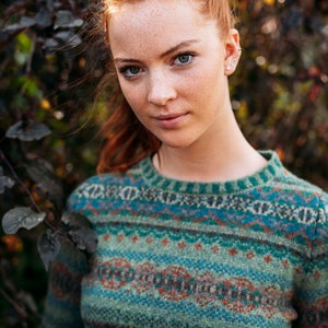 Pode incluir: Uma mulher com cabelo ruivo vestindo um suéter com padrão verde, azul e vermelho. O suéter tem um design de estilo Fair Isle.