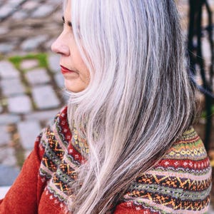 Puede incluir: Una mujer con el pelo largo, plateado y gris, lleva un suéter rojo con un patrón geométrico colorido.