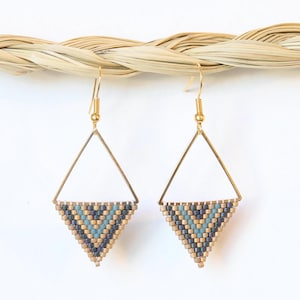 May include: Two gold-coloured, geometric earrings with a triangle shape. Each earring has a beaded design in shades of blue, grey, and tan at the bottom. The earrings are suspended against a white backdrop.