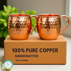 Puede incluir: Dos tazas de cobre Moscow Mule con acabado martillado. Están sobre una caja de cartón con el texto "100% PURE COPPER HANDCRAFTED Eco-Friendly". Las tazas tienen una superficie brillante y reflectante.