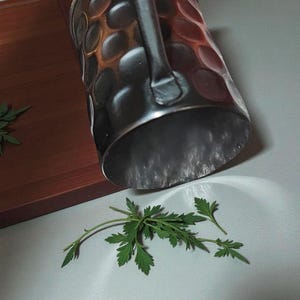 May include: A copper-colored metal mug with a handle, featuring a dimpled surface. The mug is positioned above a sprig of green leaves, with a wooden cutting board in the background. The mug is likely for beverages.