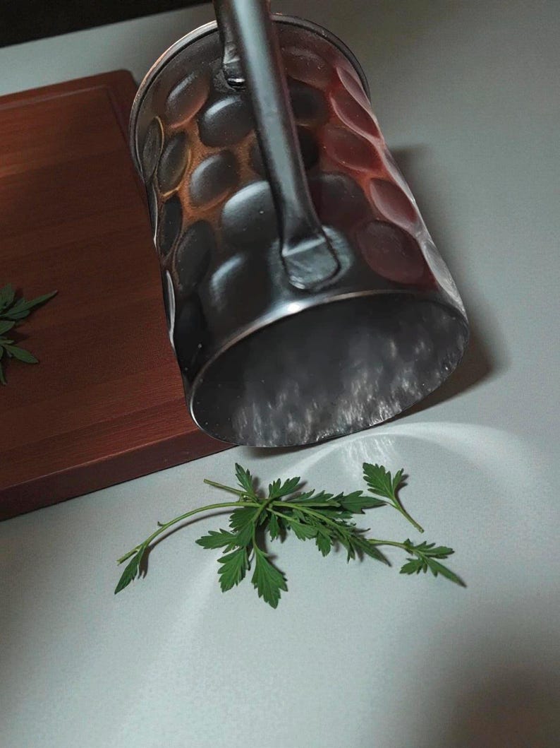 May include: A copper-colored metal mug with a handle, featuring a dimpled surface. The mug is positioned above a sprig of green leaves, with a wooden cutting board in the background. The mug is likely for beverages.