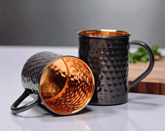 May include: Two black Moscow Mule mugs with a hammered texture. One mug is upright, the other is on its side, revealing a copper-colored interior. The mugs have a dark handle and a copper-colored rim.