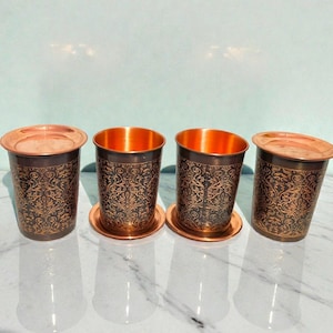 Puede incluir: Cuatro vasos de cobre con intrincados patrones florales oscuros. Cada vaso tiene un posavasos a juego y un interior de color cobre. Los vasos se exhiben sobre una superficie reflectante.