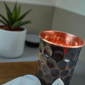 May include: A copper-colored drinking glass with a textured, dimpled exterior. The interior of the glass has a bright copper finish. A small white cloth is draped near the base of the glass. A small potted plant is in the background.