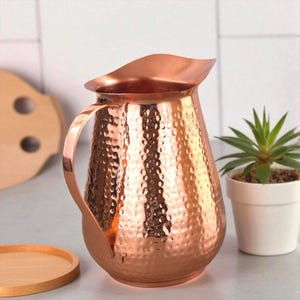 May include: A copper-coloured jug with a hammered texture. The jug has a curved handle and a spout. A small succulent plant in a white pot is to the right. A wooden serving tray is in the background.