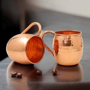 May include: Two copper Moscow Mule mugs with a hammered finish and curved handles. One mug is on its side, revealing its interior. The other mug is upright, showing its open top. Several brown coffee beans are scattered nearby.