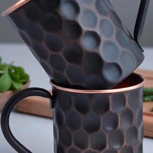 以下が含まれることがあります： 2つの銅製モスコミュールマグが積み重ねられています。マグは、暗いハンマー仕上げで、銅色の縁と黒いハンドルが付いています。マグの内側は銅色です。マグは冷たい飲み物を提供するように設計されています。