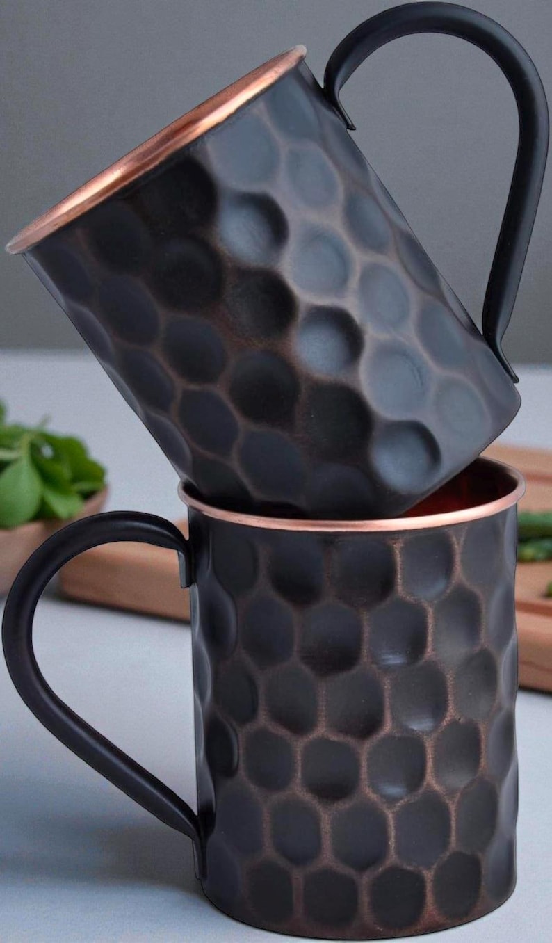 May include: Two copper Moscow Mule mugs stacked. The mugs have a dark, hammered finish with a copper rim and a black handle. The interior of the mugs is copper-colored. The mugs are designed for serving cold beverages.