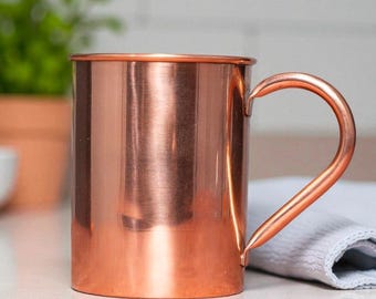 May include: A copper-colored mug with a curved handle. The mug has a cylindrical shape and a shiny, reflective surface. The handle is attached to the side of the mug and curves upwards. The mug is sitting on a white surface.