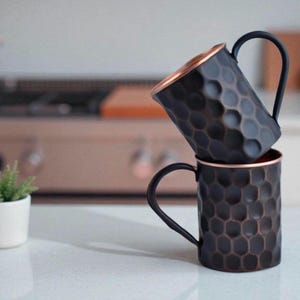 May include: Two dark copper mugs with a honeycomb pattern are stacked on a white countertop. The mugs have black handles and copper-colored interiors. The background is blurred, showing a kitchen setting with a stove and a small potted plant.