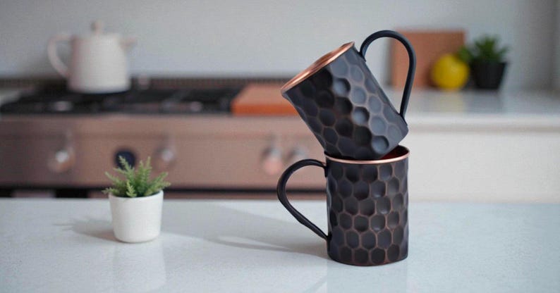 May include: Two dark copper mugs with a honeycomb pattern are stacked on a white countertop. The mugs have black handles and copper-colored interiors. The background is blurred, showing a kitchen setting with a stove and a small potted plant.