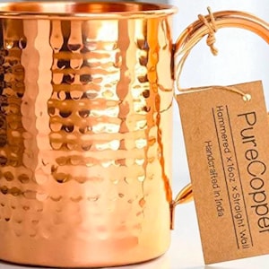 May include: A copper mug with a hammered finish and a straight wall design. The mug features a large handle and a textured surface. A tag attached to the handle reads "Pure Copper" and "Handcrafted in India". The mug is approximately 473 ml.