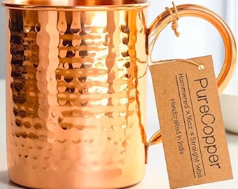 May include: A hammered copper mug with a straight wall design. The mug has a large handle and a textured surface. A tag attached to the handle reads "Pure Copper" and "Handcrafted in India". The mug is approximately 16 ounces.