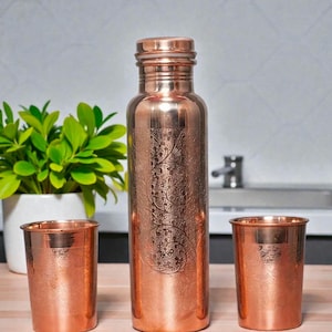 Op de afbeelding: Een koperen waterfles en twee bijpassende bekers. De fles is lang en cilindrisch met een schroefdop. De bekers zijn kort en rond. Alle items hebben een geëtst bloemmotief. Een groene plant in een witte pot staat op de achtergrond.