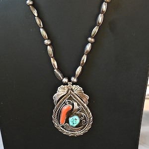 May include: A silver necklace with a pendant featuring a red coral and turquoise stone inlay. The pendant is surrounded by a decorative silver frame. The necklace has a beaded design.
