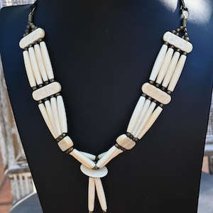 Puede incluir: Collar color crema con diseño tribal. El collar presenta múltiples hilos de cuentas cilíndricas y acentos planos y rectangulares. Las cuentas están ensartadas con un cordón negro y herrajes metálicos. El collar se exhibe en un maniquí negro.