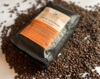 Puede incluir: Una bolsa negra de café en grano entero etiquetada como "Coffee 4 You Signature Coffee Blend" con un tueste medio y una fuerza de 4. La bolsa está sobre una superficie blanca con granos de café esparcidos alrededor.