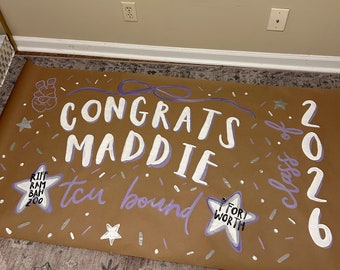 May include: A brown poster with white and purple lettering that reads "Congrats Maddie" and "TCU Bound". The poster also includes the year "2026" and the words "Fort Worth". Decorated with stars and confetti.