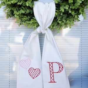 以下が含まれることがあります： 2つの刺繍されたハートと赤い文字「P」が付いた白い布。布は緑のリースに結ばれています。ハートはピンクと赤で、「P」の文字は装飾的な模様です。