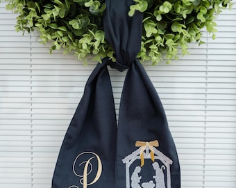 Könnte beinhalten: Ein Weihnachtskranz mit einem dunkelblauen Schal mit einer gestickten Krippenszene und dem Buchstaben "P". Der Kranz besteht aus grünen Blättern und hängt vor einem weißen Hintergrund.