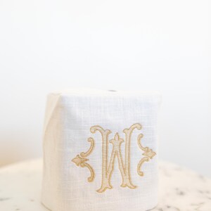 May include: A white linen tissue box cover with an embroidered gold letter "W" in an ornate script. The cover is square and sits on a marble-topped table, offering a touch of elegance to any bathroom or bedroom.