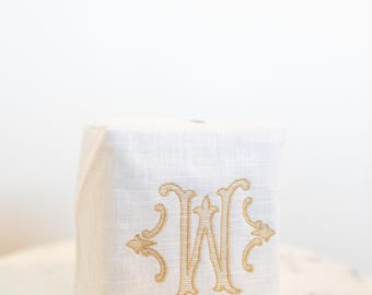Könnte beinhalten: Eine weiße Tissuebox-Hülle aus Leinen mit einem gestickten goldenen Buchstaben "W" in einer verzierten Schrift. Die quadratische Hülle steht auf einem Tisch mit Marmorplatte und verleiht jedem Badezimmer oder Schlafzimmer einen Hauch von Eleganz.