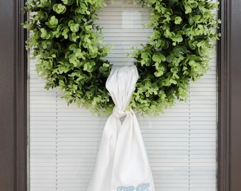May include: A green leafy wreath hangs on a door with white blinds. A white cloth with a light blue embroidered letter "V" is tied to the wreath. The wreath is a full circle of green leaves.