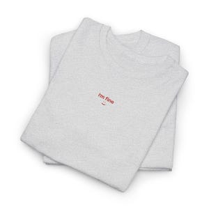 以下が含まれることがあります： 2枚のライトグレーのTシャツが重ねられています。上のTシャツには、赤い糸で「I'm fine」の文字と小さな笑顔が刺繍されています。Tシャツはきれいに折りたたまれています。シンプルでカジュアルなデザインです。