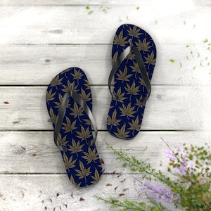May include: Navy blue flip-flops featuring a repeating gold cannabis leaf design. The sandals have black straps and are set against a white wooden backdrop. The pattern is identical on both flip-flops, creating a matching set.