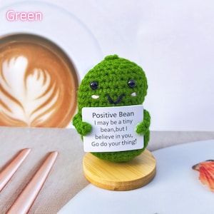 May include: Green crocheted bean plush holding a sign that reads "Positive Bean I may be a tiny bean, but I believe in you, Go do your thing!"