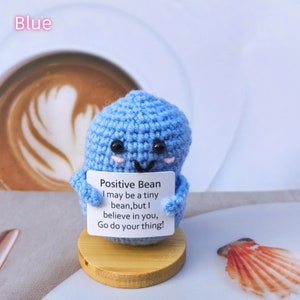 May include: A blue crocheted bean-shaped plushie holding a sign that reads "Positive Bean I may be a tiny bean, but I believe in you, Go do your thing!"
