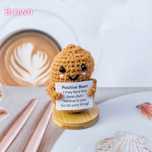 May include: A small crocheted bean-shaped figure holding a sign that reads "Positive Bean I may be a tiny bean, but I believe in you, Go do your thing!"