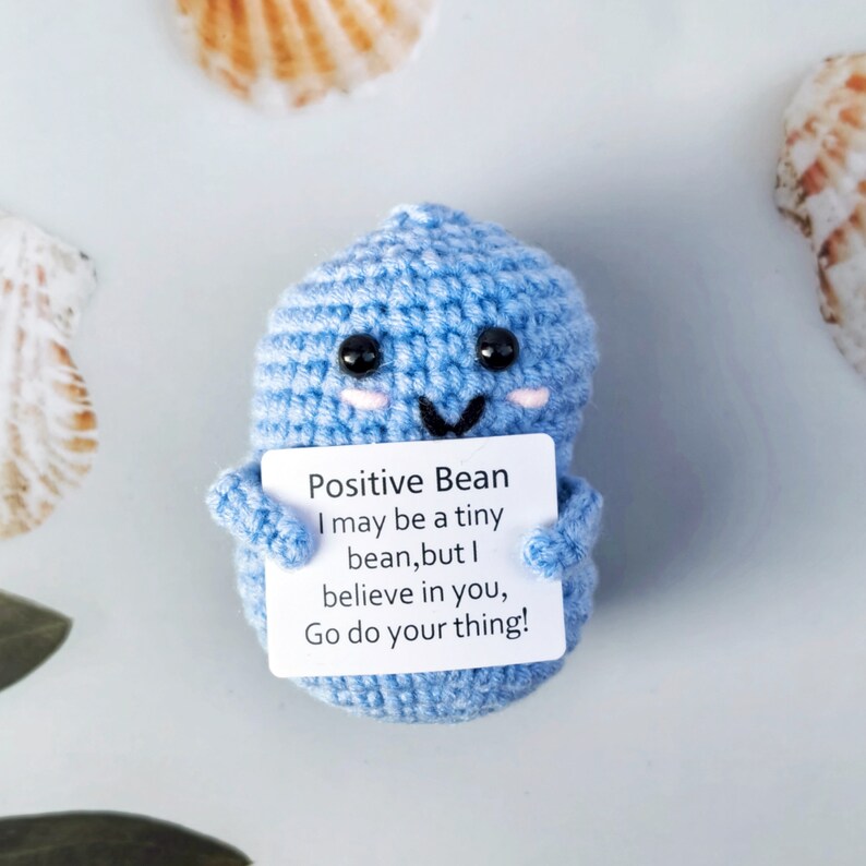 May include: A blue crocheted bean-shaped plush toy holding a card that reads "Positive Bean I may be a tiny bean, but I believe in you, Go do your thing!"