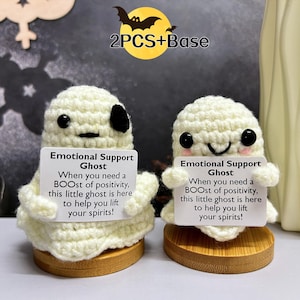 Könnte beinhalten: Zwei gehäkelte weiße Geisterfiguren, die Schilder mit der Aufschrift "Emotional Support Ghost" halten, mit dem Text "When you need a BOOST of positivity, this little ghost is here to help you lift your spirits!" Die Geister sitzen auf kleinen Holzsockeln.
