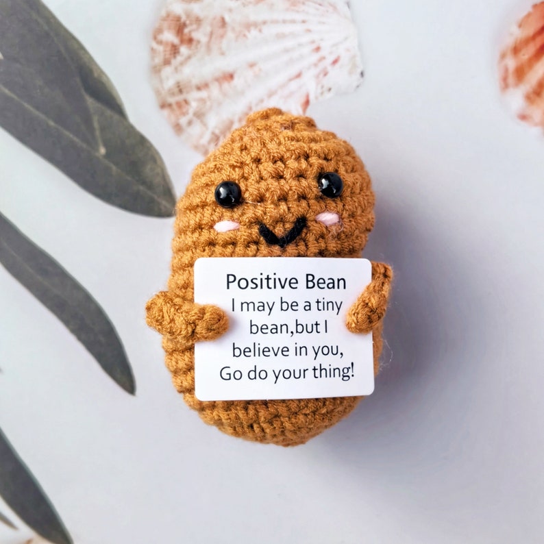 May include: A crocheted bean-shaped plush toy holding a sign that reads "Positive Bean I may be a tiny bean, but I believe in you, Go do your thing!"