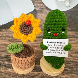 May include: A crocheted sunflower with a ladybug on the center, and a crocheted green pickle holding a sign that reads "Positive Pickle I may be a tiny pickle, but I believe in you, go do your things!"