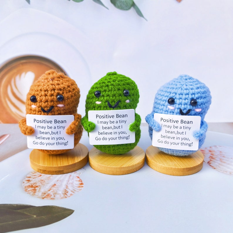 May include: Three crocheted bean plushies, each holding a sign with the text "Positive Bean I may be a tiny bean, but I believe in you, Go do your thing!" The beans are brown, green, and blue.