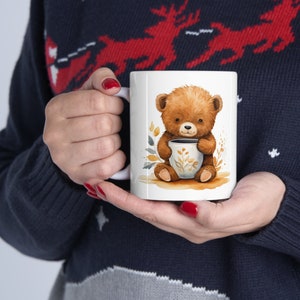 Könnte beinhalten: Weiße Keramiktasse mit einer Aquarellillustration eines braunen Teddybären, der eine Tasse mit floralem Design hält. Die Tasse hat einen weißen Henkel. Die Person, die die Tasse hält, trägt einen marineblauen Pullover.