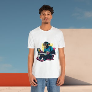 May include: White t-shirt with a vibrant graphic of a chimpanzee using a laptop. The chimpanzee is wearing a headband and a blue shirt. The background is a mix of blue and white. The design is eye-catching.